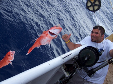 Chris Monsour smiles and gives a thumbs up while on a research ship, standing near a fishing line with two bright orange fish over deep blue ocean water.