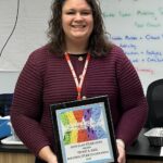 A woman with curly hair wearing a maroon sweater stands indoors, smiling, and holding a framed award that reads "Rhode Island STEAM Center High School STEAM Educator Award 2025."