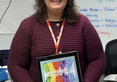 A woman with curly hair wearing a maroon sweater stands indoors, smiling, and holding a framed award that reads "Rhode Island STEAM Center High School STEAM Educator Award 2025."