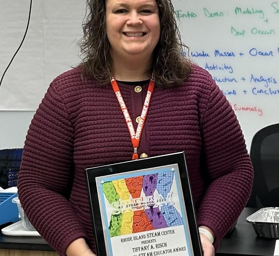 A woman with curly hair wearing a maroon sweater stands indoors, smiling, and holding a framed award that reads "Rhode Island STEAM Center High School STEAM Educator Award 2025."