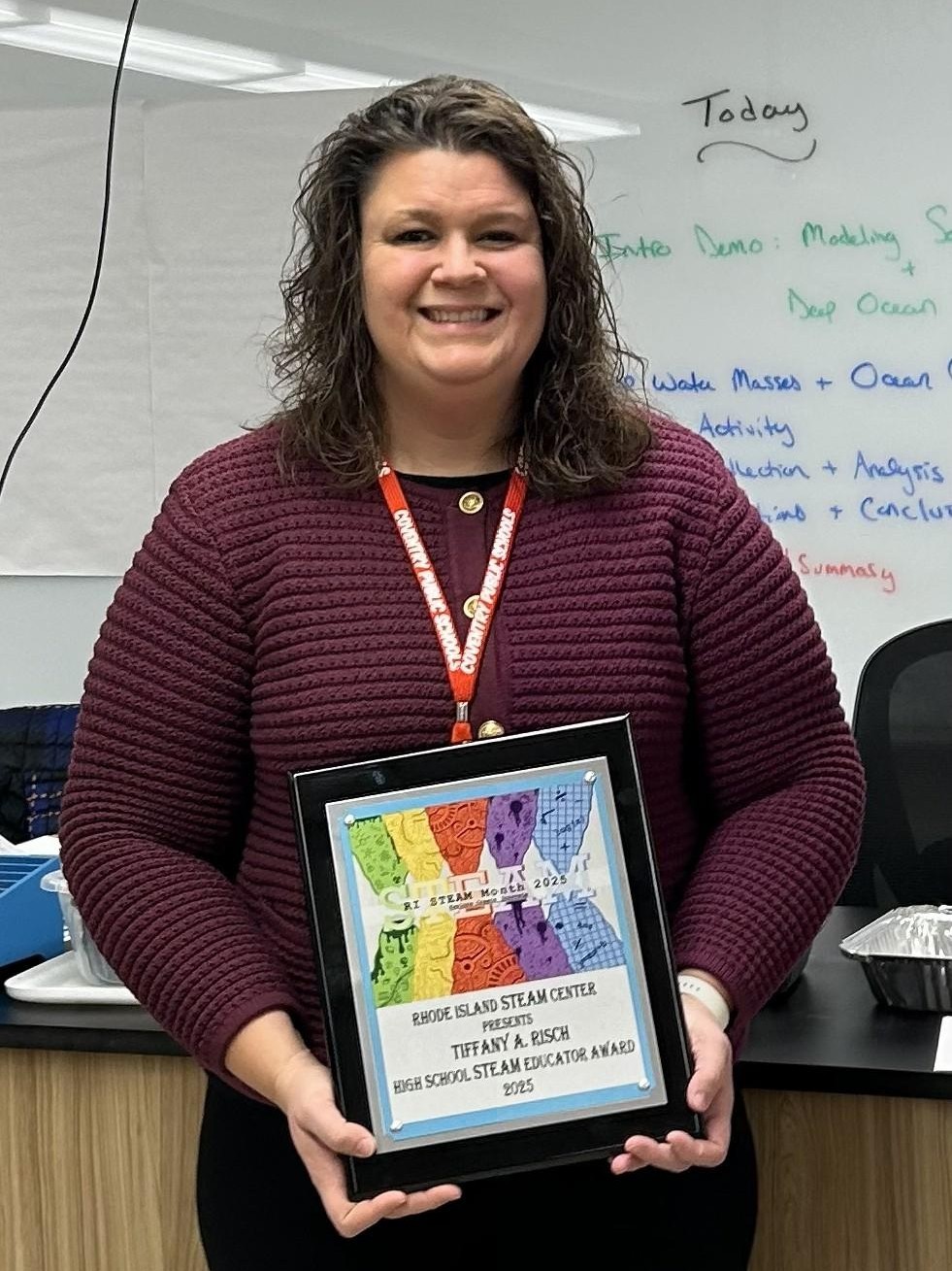 A woman with curly hair wearing a maroon sweater stands indoors, smiling, and holding a framed award that reads "Rhode Island STEAM Center High School STEAM Educator Award 2025."