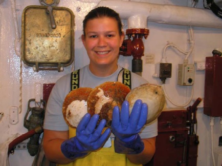 Tiffany Risch, a 2008 NOAA Teacher at Sea, smiles while wearing blue gloves and yellow overalls, holding three large sea scallops inside a ship laboratory lined with pipes and equipment.