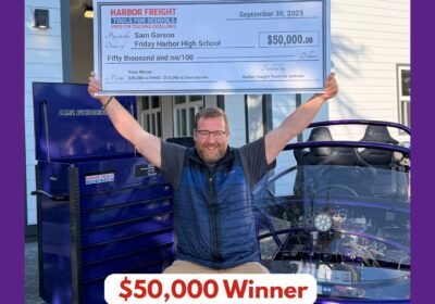A man smiles and raises his arms in victory while holding a large $50,000 STEAM Educator Award check from Harbor Freight Tools for Schools outside Friday Harbor High School, next to a blue vehicle.
