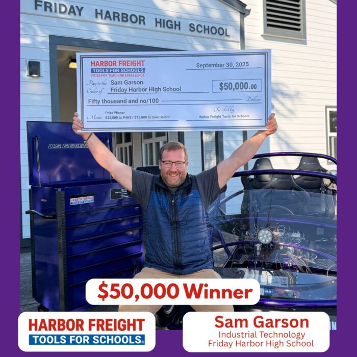 A man smiles and raises his arms in victory while holding a large $50,000 STEAM Educator Award check from Harbor Freight Tools for Schools outside Friday Harbor High School, next to a blue vehicle.