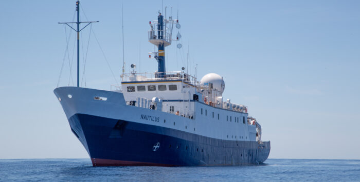 The large research vessel E/V Nautilus floats on calm blue water under a clear sky, antennas and equipment lining the upper deck.
