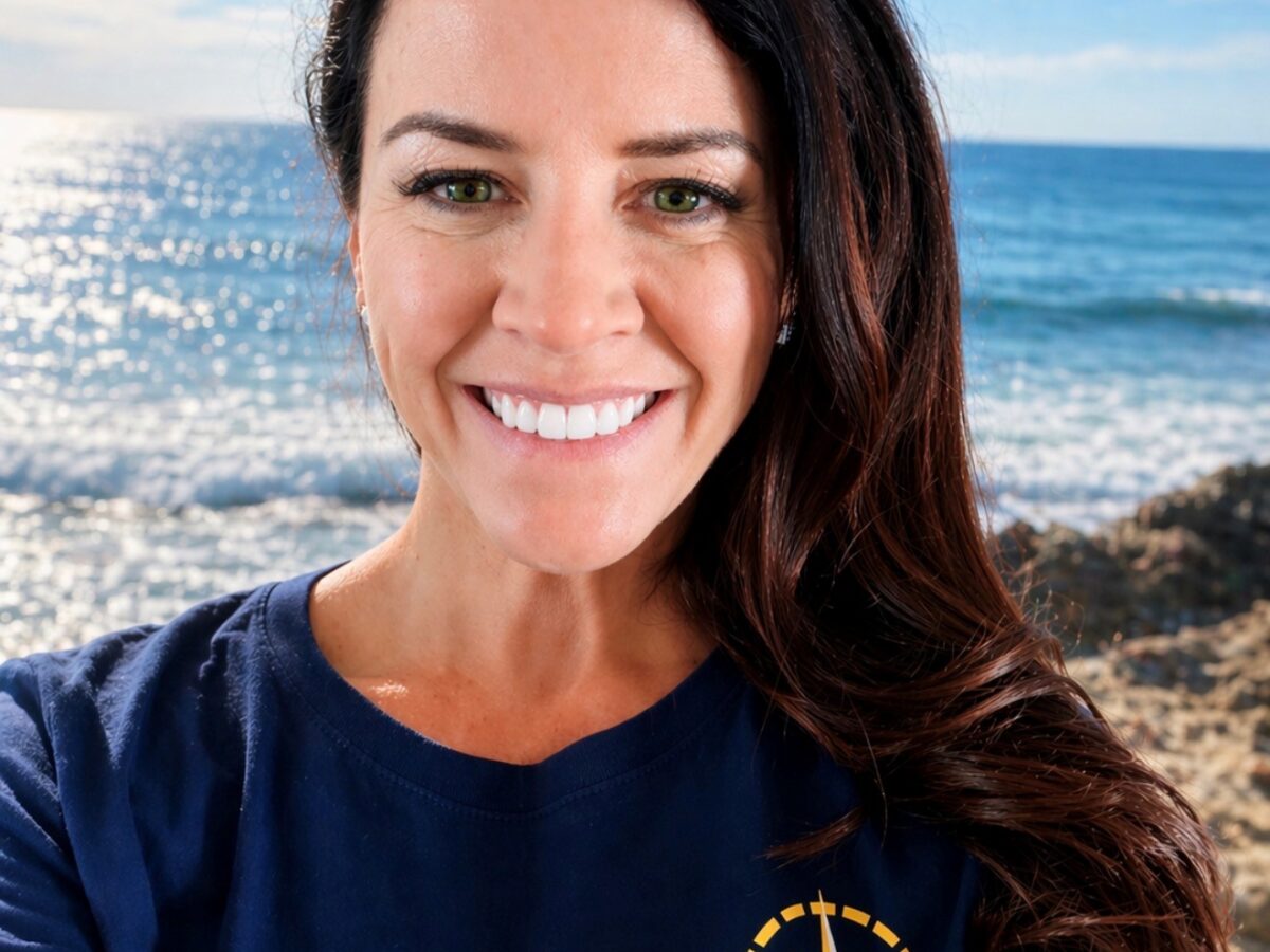 A smiling woman with long dark hair, a NOAA Teacher at Sea, wears a navy "E/V Nautilus Ocean Exploration Trust" t-shirt and stands in front of a sunny ocean view with blue sky, waves, and rocky shore.