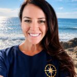 A smiling woman with long dark hair, a NOAA Teacher at Sea, wears a navy "E/V Nautilus Ocean Exploration Trust" t-shirt and stands in front of a sunny ocean view with blue sky, waves, and rocky shore.