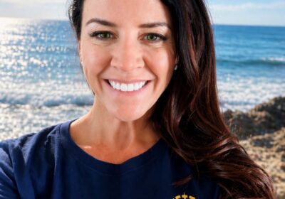 A smiling woman with long dark hair, a NOAA Teacher at Sea, wears a navy "E/V Nautilus Ocean Exploration Trust" t-shirt and stands in front of a sunny ocean view with blue sky, waves, and rocky shore.