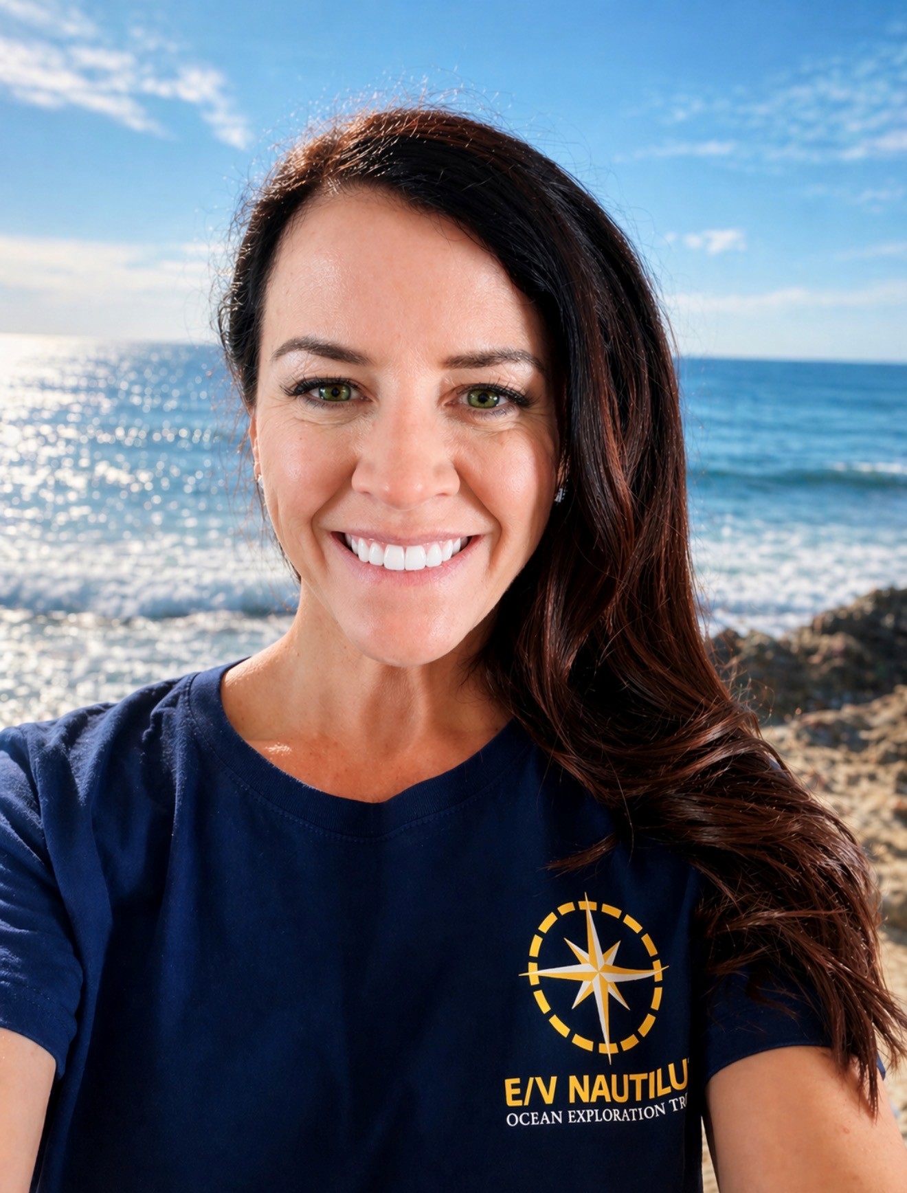 A smiling woman with long dark hair, a NOAA Teacher at Sea, wears a navy "E/V Nautilus Ocean Exploration Trust" t-shirt and stands in front of a sunny ocean view with blue sky, waves, and rocky shore.