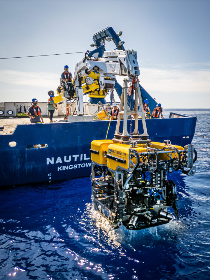 A yellow remotely operated underwater vehicle is being lowered into blue ocean water from the E/V Nautilus as crew members in safety gear work on deck under a clear sky, supporting Pacific exploration.